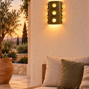 Op de afbeelding: Een cilindervormige olijfgroene wandlamp met zes ronde uitsparingen in twee verticale kolommen. Het armatuur verspreidt een warm, gloeiend licht en werpt een lichtbundel naar boven en beneden op het oppervlak van de muur.