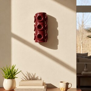 Op de afbeelding: Een cilindrische wandlamp van donkerbordeaux keramiek met een herhalend patroon van verhoogde, cirkelvormige ringmotieven over het verticale oppervlak. Het object is bevestigd aan een vlakke muur en werpt een schaduw.