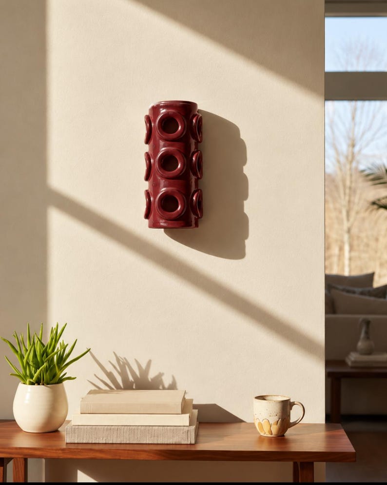 Op de afbeelding: Een cilindrische wandlamp van donkerbordeaux keramiek met een herhalend patroon van verhoogde, cirkelvormige ringmotieven over het verticale oppervlak. Het object is bevestigd aan een vlakke muur en werpt een schaduw.
