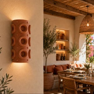 Op de afbeelding: Een cilindervormige wandlamp van terracotta met een herhalend patroon van cirkelvormige inkepingen en kleine verhoogde stippen. Het armatuur verspreidt een warme, zachte gloed aan de boven- en onderkant op de beige muur.