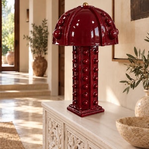 Puede incluir: Una lámpara de mesa de cerámica de color rojo oscuro brillante con una pantalla en forma de hongo y una base cuadrada, ambas decoradas con filas de tachuelas esféricas en relieve. La lámpara tiene un remate esférico dorado.