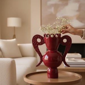 May include: A glossy, deep red decorative vase with two ornate handles and a scalloped rim. The vase is filled with white flowers and sits on a round wooden table. The vase is a striking home decor accent.