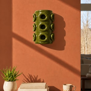 Op de afbeelding: Een cilindrische olijfgroene keramische wandplantenbak met een herhalend patroon van verhoogde cirkelvormige openingen in drie verticale rijen. Het glanzende object met een glad oppervlak is bevestigd aan een vlakke muur.