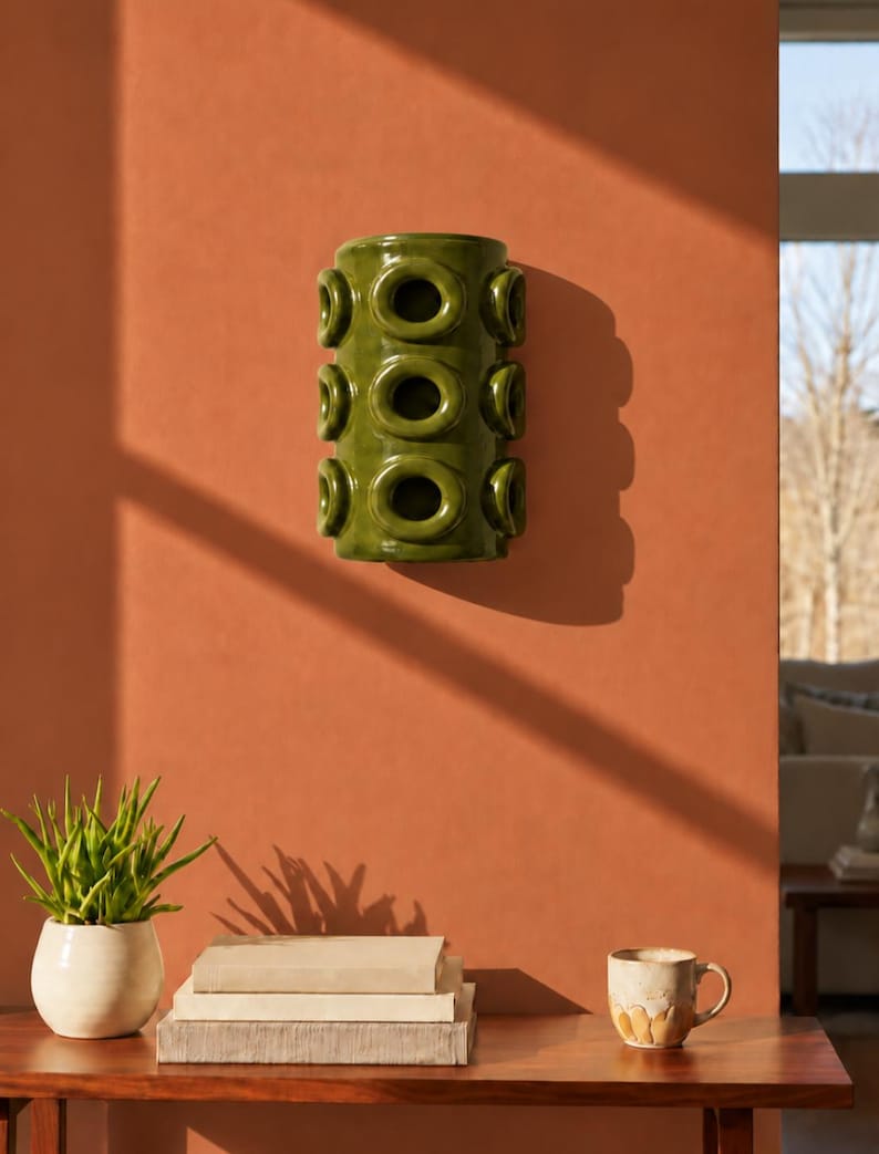 Op de afbeelding: Een cilindrische olijfgroene keramische wandplantenbak met een herhalend patroon van verhoogde cirkelvormige openingen in drie verticale rijen. Het glanzende object met een glad oppervlak is bevestigd aan een vlakke muur.