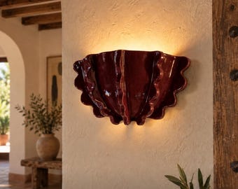 Op de afbeelding: Een halfronde wandlamp met een diepe bordeauxrode, glanzende keramische afwerking. Het armatuur heeft een geschulpt, schelpachtig ontwerp met verticale ribbels die naar buiten stralen en een warme, zachte gloed verspreiden.