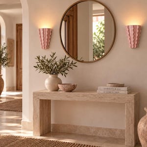 Puede incluir: Una mesa consola de travertino beige sostiene un jarr&oacute;n de cer&aacute;mica blanca con ramas verdes, un cuenco peque&ntilde;o y dos libros titulados The Kinfolk Home y At Home in Spain. Un espejo redondo est&aacute; flanqueado por dos apliques rosas.