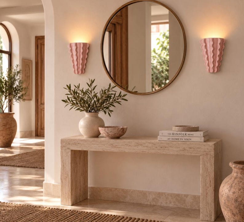 Puede incluir: Una mesa consola de travertino beige sostiene un jarr&oacute;n de cer&aacute;mica blanca con ramas verdes, un cuenco peque&ntilde;o y dos libros titulados The Kinfolk Home y At Home in Spain. Un espejo redondo est&aacute; flanqueado por dos apliques rosas.