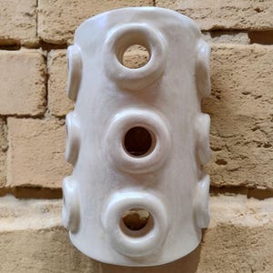 Op de afbeelding: Een cilindervormig, gebroken wit keramieken vat met een reeks verhoogde, cirkelvormige openingen in een verticaal patroon op het buitenoppervlak. Het object heeft een gladde, glanzende afwerking en een holle kern met ringvormige uitsteeksels.