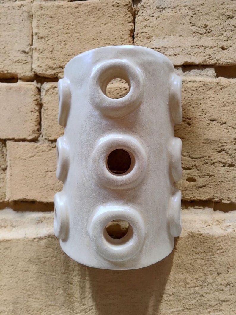Op de afbeelding: Een cilindervormig, gebroken wit keramieken vat met een reeks verhoogde, cirkelvormige openingen in een verticaal patroon op het buitenoppervlak. Het object heeft een gladde, glanzende afwerking en een holle kern met ringvormige uitsteeksels.