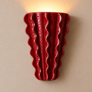 Puede incluir: Un aplique de pared de cer&aacute;mica en color rojo intenso brillante con una silueta vertical en forma de abanico, que presenta crestas onduladas prominentes que se extienden desde la base estrecha hasta la parte superior, emitiendo luz.