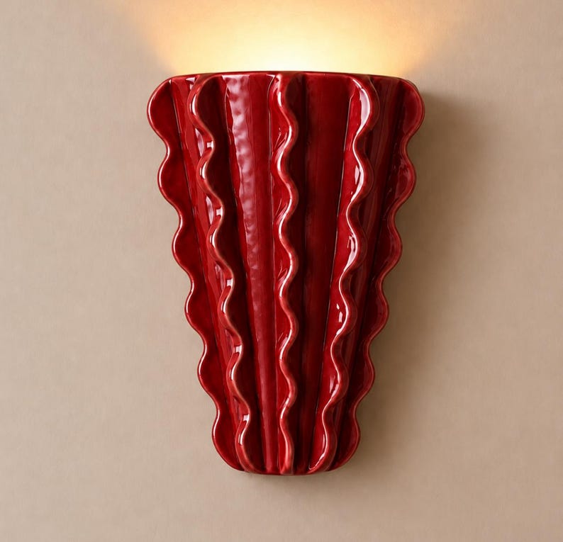 Puede incluir: Un aplique de pared de cer&aacute;mica en color rojo intenso brillante con una silueta vertical en forma de abanico, que presenta crestas onduladas prominentes que se extienden desde la base estrecha hasta la parte superior, emitiendo luz.