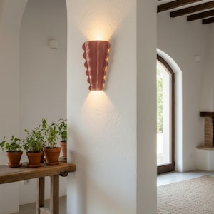 Puede incluir: Un aplique de pared con una pantalla de cer&aacute;mica brillante de color rosa empolvado, con un dise&ntilde;o de bordes verticales festoneados u ondulados. La l&aacute;mpara emite un brillo c&aacute;lido y suave desde las aberturas superior e inferior.