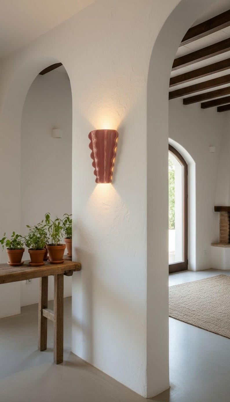 Puede incluir: Un aplique de pared con una pantalla de cer&aacute;mica brillante de color rosa empolvado, con un dise&ntilde;o de bordes verticales festoneados u ondulados. La l&aacute;mpara emite un brillo c&aacute;lido y suave desde las aberturas superior e inferior.