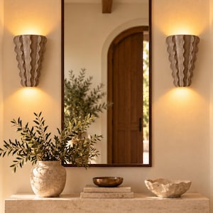 Puede incluir: Un espejo rectangular con marco oscuro, flanqueado por dos apliques de pared beige con estr&iacute;as verticales que emiten luz c&aacute;lida. Debajo, una consola de piedra sostiene un jarr&oacute;n beige texturizado, un cuenco de bronce y un cuenco de piedra.