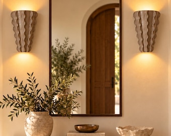 Op de afbeelding: Een rechthoekige wandspiegel met een donker frame, geflankeerd door twee beige, verticaal gegroefde wandlampen die warm licht uitstralen. Op de stenen console eronder staan een beige vaas met textuur, een kleine bronzen schaal en een stenen schaal.