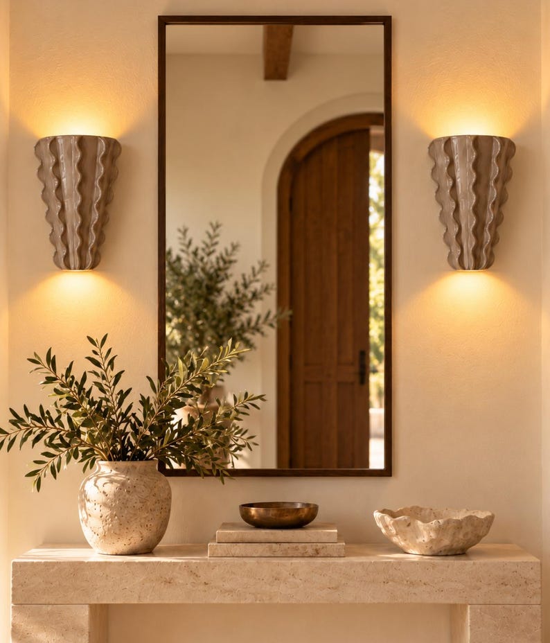 Puede incluir: Un espejo rectangular con marco oscuro, flanqueado por dos apliques de pared beige con estr&iacute;as verticales que emiten luz c&aacute;lida. Debajo, una consola de piedra sostiene un jarr&oacute;n beige texturizado, un cuenco de bronce y un cuenco de piedra.