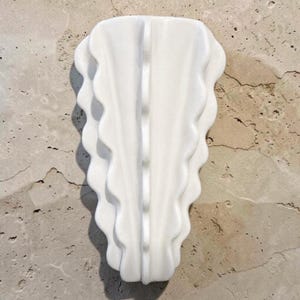 Puede incluir: Un florero de pared de cer&aacute;mica blanca con una cresta central vertical y bordes festoneados en capas que se abren sim&eacute;tricamente. El objeto tiene un acabado liso y brillante, con una forma alargada y c&oacute;nica para su montaje.