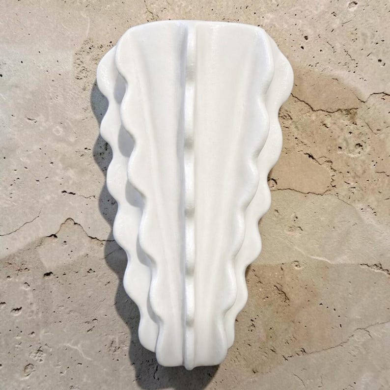Puede incluir: Un florero de pared de cer&aacute;mica blanca con una cresta central vertical y bordes festoneados en capas que se abren sim&eacute;tricamente. El objeto tiene un acabado liso y brillante, con una forma alargada y c&oacute;nica para su montaje.