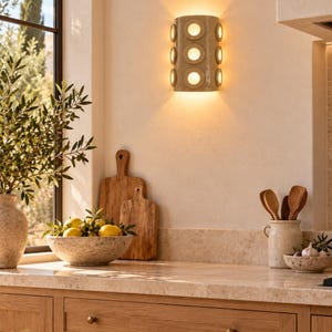 Op de afbeelding: Een halfcilindervormige wandlamp met een glanzende beige afwerking en zes cirkelvormige uitsparingen in twee verticale kolommen, die warm, diffuus licht verspreidt tegen een neutrale keukenmuur.