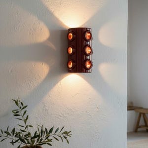 Op de afbeelding: Een cilindervormige wandlamp met een glanzende donkerbruine afwerking en zes ronde openingen in twee verticale kolommen. Het armatuur verspreidt warm licht vanaf de bovenkant, onderkant en de zes ronde poorten, wat opvallende lichtpatronen op de muur werpt.