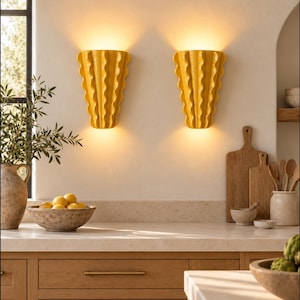 Puede incluir: Dos apliques de pared de cer&aacute;mica amarilla con un dise&ntilde;o vertical ondulado y plisado. Cada aplique emite un brillo c&aacute;lido y suave desde la parte superior e inferior, iluminando la pared. Las l&aacute;mparas tienen un acabado brillante y forma de abanico.