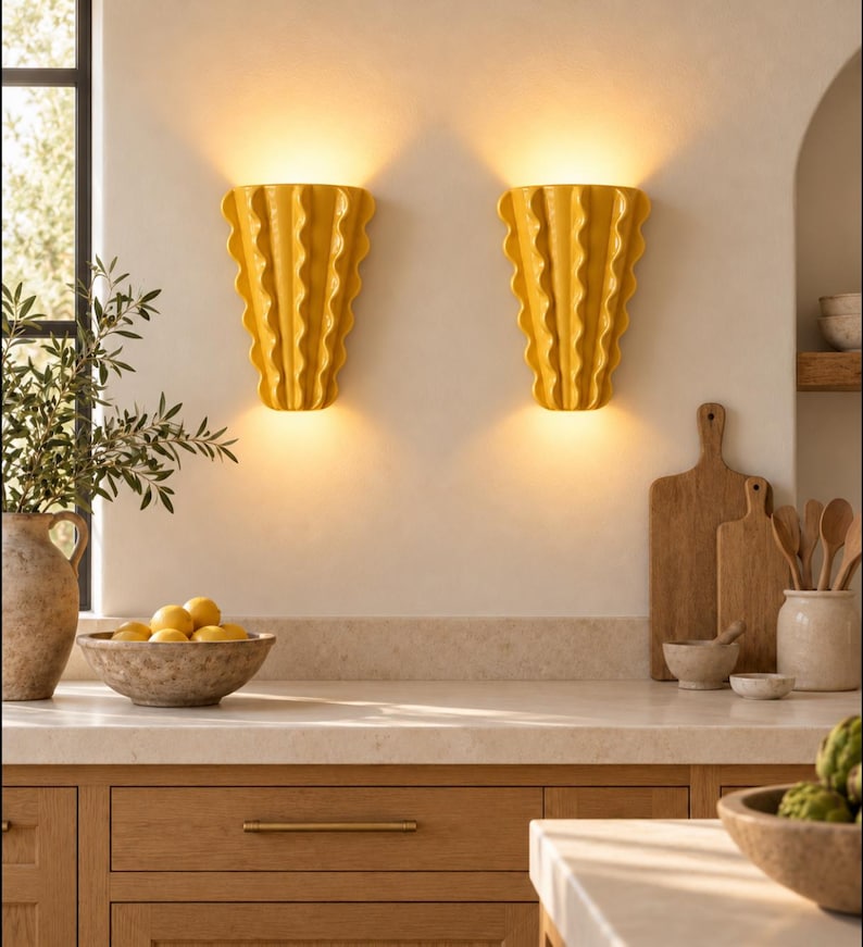 Puede incluir: Dos apliques de pared de cer&aacute;mica amarilla con un dise&ntilde;o vertical ondulado y plisado. Cada aplique emite un brillo c&aacute;lido y suave desde la parte superior e inferior, iluminando la pared. Las l&aacute;mparas tienen un acabado brillante y forma de abanico.