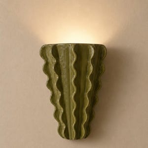Puede incluir: Un aplique de pared con un cuerpo de cer&aacute;mica verde oliva brillante y una silueta vertical ondulada en forma de abanico. La l&aacute;mpara emite un brillo suave y c&aacute;lido desde la parte superior, proyectando luz hacia arriba sobre la pared.