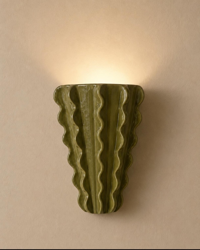 Puede incluir: Un aplique de pared con un cuerpo de cer&aacute;mica verde oliva brillante y una silueta vertical ondulada en forma de abanico. La l&aacute;mpara emite un brillo suave y c&aacute;lido desde la parte superior, proyectando luz hacia arriba sobre la pared.