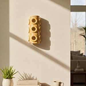Op de afbeelding: Een cilindervormige gele keramische wandlamp met een herhalend patroon van verhoogde cirkelvormige ringen aan de buitenkant. Het armatuur is verticaal op een effen muur gemonteerd en werpt een duidelijke schaduw aan de rechterkant.