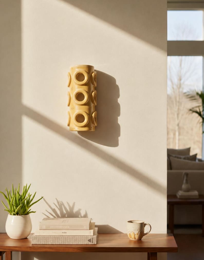Op de afbeelding: Een cilindervormige gele keramische wandlamp met een herhalend patroon van verhoogde cirkelvormige ringen aan de buitenkant. Het armatuur is verticaal op een effen muur gemonteerd en werpt een duidelijke schaduw aan de rechterkant.