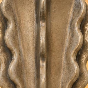 Puede incluir: Un objeto de cer&aacute;mica en tono bronce met&aacute;lico que presenta una cresta vertical central flanqueada por curvas sim&eacute;tricas y onduladas. La superficie tiene un acabado texturizado y desgastado con variaciones de luz y sombra.
