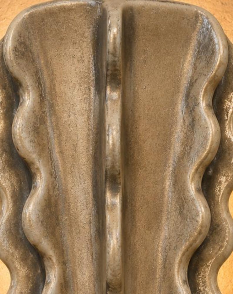 Puede incluir: Un objeto de cer&aacute;mica en tono bronce met&aacute;lico que presenta una cresta vertical central flanqueada por curvas sim&eacute;tricas y onduladas. La superficie tiene un acabado texturizado y desgastado con variaciones de luz y sombra.