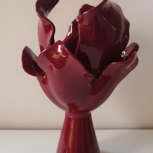 Może przedstawiać: Ciemnoczerwony wazon ceramiczny z błyszczącym wykończeniem, o rzeźbiarskim kształcie przypominającym otwarty kwiat. Podstawa jest wąska i cylindryczna, rozszerzająca się ku górze w duże, zakrzywione formy przypominające płatki kwiatów.