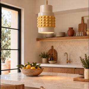 May include: A cylindrical pendant light fixture featuring a smooth cream-colored top and bottom section with a central band of raised, rounded yellow spheres. The light hangs from a thin metal rod and provides downward illumination.