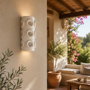 Op de afbeelding: Een cilindervormige witte keramische wandlamp met drie verticaal geplaatste ronde uitsparingen. Het armatuur verspreidt een warm licht vanuit de bovenste en onderste openingen op de getextureerde beige muur.