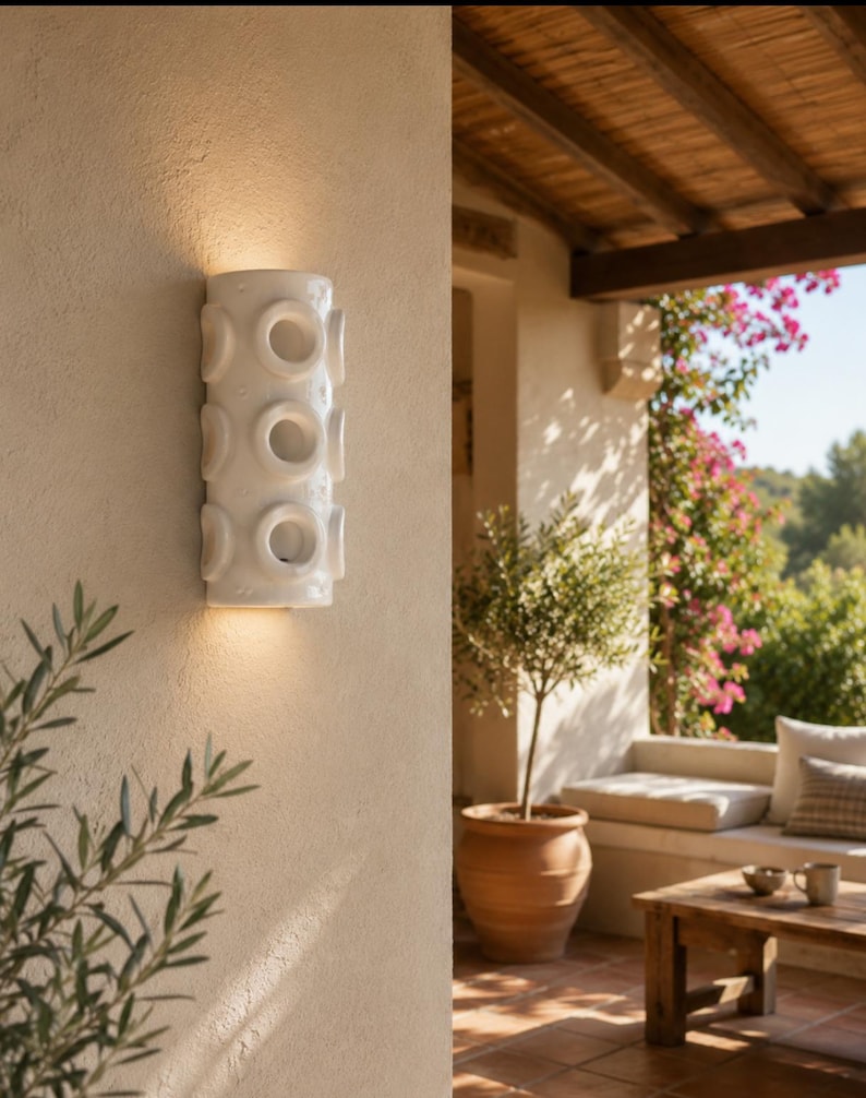 Op de afbeelding: Een cilindervormige witte keramische wandlamp met drie verticaal geplaatste ronde uitsparingen. Het armatuur verspreidt een warm licht vanuit de bovenste en onderste openingen op de getextureerde beige muur.