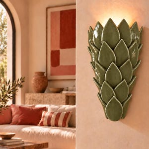 Puede incluir: Un aplique de pared con un diseño de hojas superpuestas en un acabado verde oliva brillante. La lámpara está montada en una pared de color claro y emite un brillo cálido y suave desde la parte superior, resaltando la estructura cerámica texturizada.