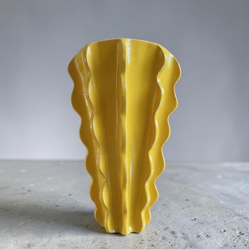 Puede incluir: Un jarr&oacute;n de cer&aacute;mica de color amarillo brillante con una estructura vertical, ondulada y plisada. El objeto presenta una abertura ancha en forma de abanico en la parte superior que se estrecha hacia una base delgada, creando pliegues.
