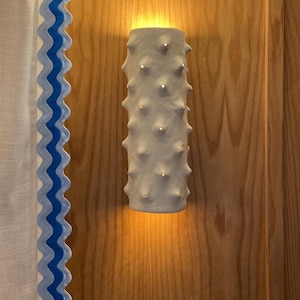 May include: A white, cylindrical wall sconce with a textured, spiky surface is mounted on a wooden wall. The sconce emits a warm light. A white curtain with a blue wavy trim is on the left.