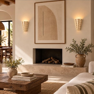 Puede incluir: Una sala de estar con chimenea, dos apliques de pared estriados en color beige, un cuadro abstracto enmarcado, una mesa de centro de madera con libros apilados y dos jarrones de cerámica con ramas verdes sobre una pared neutra.