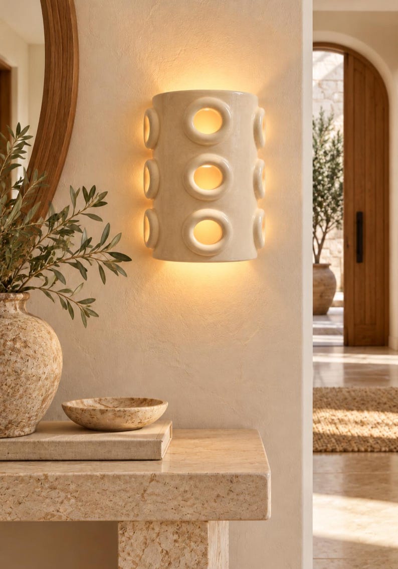 Op de afbeelding: Een halfcilindervormige wandlamp van gebroken wit keramiek, voorzien van drie verticale rijen cirkelvormige uitsparingen die een warm, gloeiend licht uitstralen. Het armatuur is vlak tegen een gestructureerde muur gemonteerd en verspreidt zacht licht.