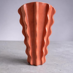 Puede incluir: Un jarr&oacute;n de cer&aacute;mica color terracota con una estructura vertical ondulada en forma de abanico. El objeto presenta un acabado brillante y un borde superior ancho y festoneado que se estrecha hacia una base estrecha y acanalada.