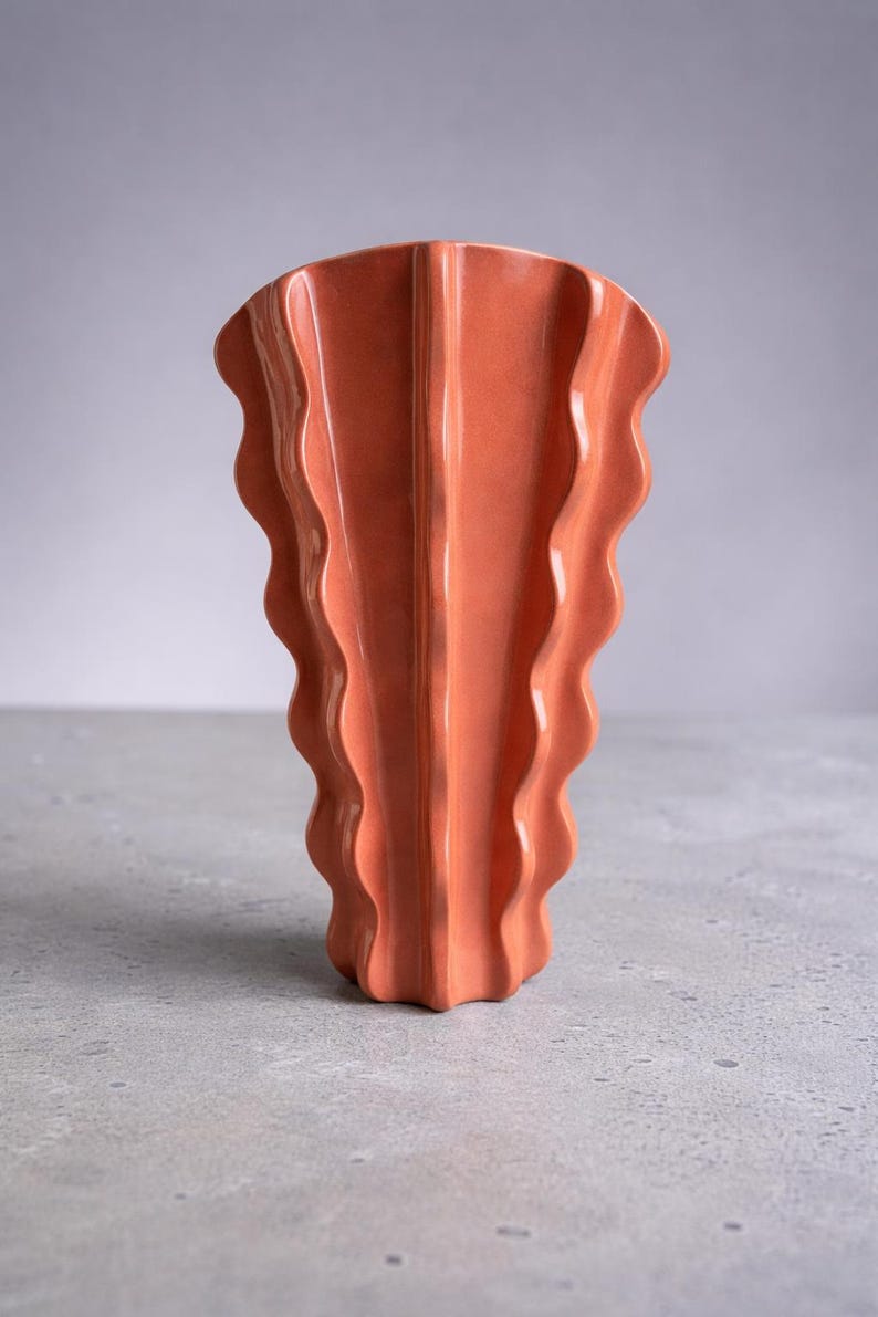 Puede incluir: Un jarr&oacute;n de cer&aacute;mica color terracota con una estructura vertical ondulada en forma de abanico. El objeto presenta un acabado brillante y un borde superior ancho y festoneado que se estrecha hacia una base estrecha y acanalada.