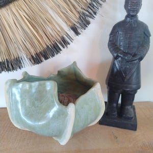 Puede incluir: Una composición decorativa con un cuenco de cerámica verde claro de forma única, una estatua oscura de un guerrero y un adorno de pared circular hecho de fibras naturales. Los objetos se exhiben sobre una superficie de madera.