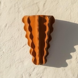 Puede incluir: Un aplique de pared de madera con un acabado c&aacute;lido en color marr&oacute;n medio. El objeto tiene una forma triangular distintiva que se estrecha hacia abajo, con bordes verticales festoneados que crean un patr&oacute;n r&iacute;tmico y ondulado en los lados.