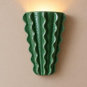 Puede incluir: Un aplique de pared con un cuerpo de cer&aacute;mica verde oscuro brillante y un dise&ntilde;o de bordes verticales ondulados y con volantes. La l&aacute;mpara emite un brillo c&aacute;lido y suave desde la parte superior, resaltando la superficie lisa y reflectante.