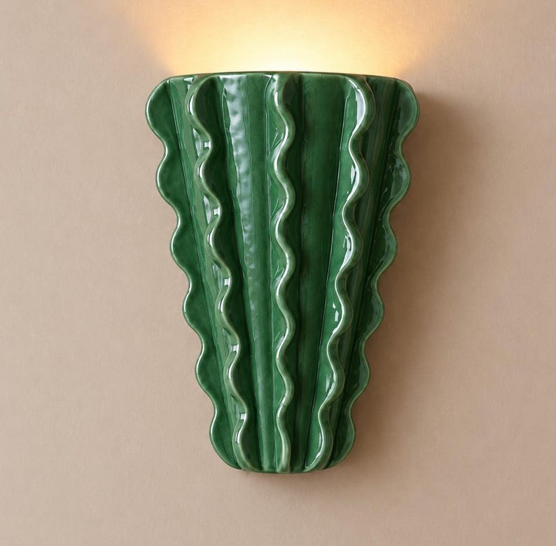 Puede incluir: Un aplique de pared con un cuerpo de cer&aacute;mica verde oscuro brillante y un dise&ntilde;o de bordes verticales ondulados y con volantes. La l&aacute;mpara emite un brillo c&aacute;lido y suave desde la parte superior, resaltando la superficie lisa y reflectante.
