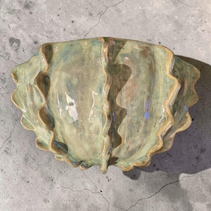 Può includere: Una ciotola in ceramica con bordo ondulato e smerlato, caratterizzata da una smaltatura lucida nei toni del verde salvia, azzurro pallido e beige. La ciotola presenta una distinta cresta verticale centrale, che le conferisce un aspetto a conchiglia.
