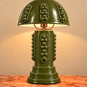 Puede incluir: Una lámpara de mesa con acabado cerámico en color verde oliva brillante, que presenta una pantalla en forma de cúpula y una base cilíndrica. Tanto la pantalla como la base están decoradas con paneles verticales de texturas esféricas en relieve.
