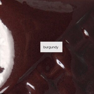 Op de afbeelding: Een glad, glanzend oppervlak met een diepe bordeauxrode kleur. Een wit rechthoekig label is in het midden van de afbeelding geplaatst, met het woord burgundy in zwarte, schreefloze kleine letters. De afwerking is reflecterend.
